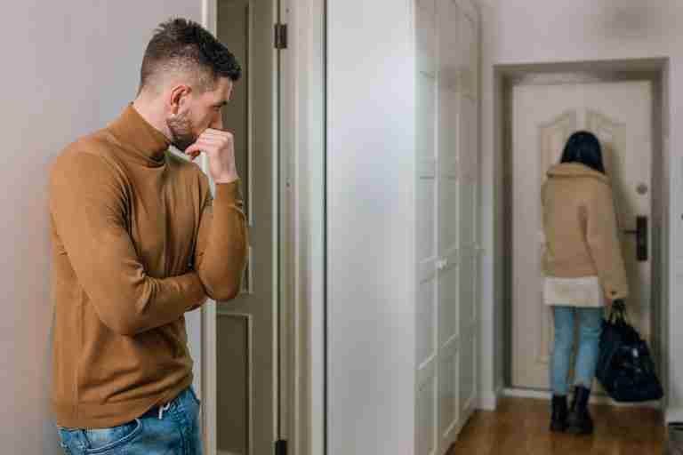 In this image, a man wearing an orange turtleneck watches around the corner as a woman with a suitcase leaves through a door to their New York City apartment. The image represents a breakup