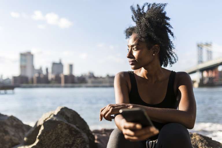 New York City, Brooklyn, woman with cell phone sitting at the waterfront looking up the power of DBT skills in treating BPD